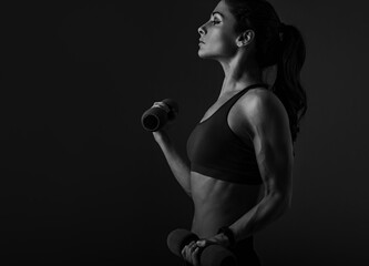 Sporty muscular serious woman doing strength workout on the shoulders, biceps and arms in sport bra holding dumbbells on grey dark shadow background with empty space. Closeup profile.