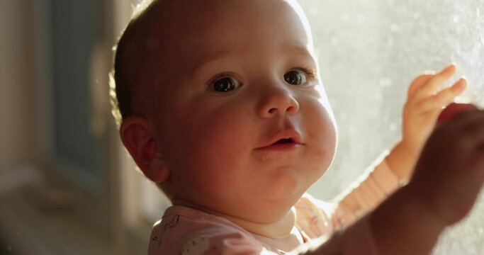 Toddler girl leaning against low window on sunny morning looking towards camera - cute golden hour look
