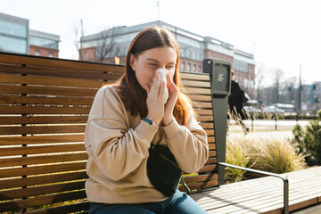 Fototapeta premium Unhappy caucasian millennial red-haired woman sneezing in napkin and siting in park bench, Young women suffering from allergy outdoors