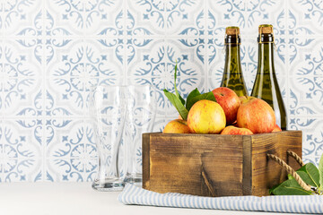Apple cider drink or fermented fruit drink. Bottles with cider, glasses and red organic apples in a wooden box on a kitchen table opposite tile wall. Healthy food. Apple, low alcohol drink
