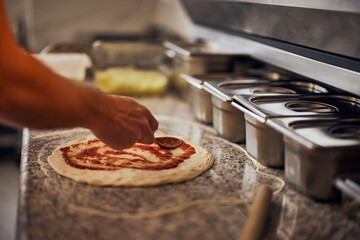 A side view of a male pizza master spreading tomato sauce on dough.