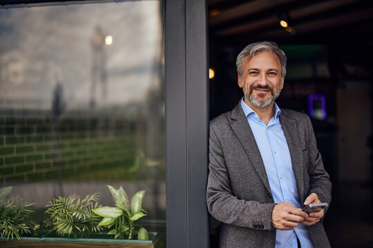 A portrait of a senior male businessman stands at the entrance of his restaurant.