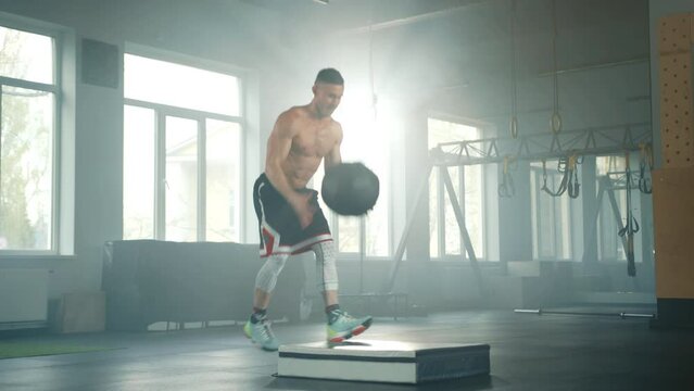 Young handsome sportsman with bare torso explosively lifts medical ball overhead, throws it on balance pad while jumping from one leg to another in gym. High quality 4k footage