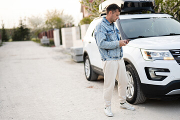 Man in jeans jacket with phone in hand standing near white american suv car with roof rack box.