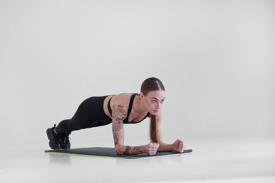 A young girl with brown hair, with a tattoo in a black top and leggings on a white background. Sport, fitness, stands in the bar. Press exercises. Yoga
