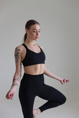 A young girl with brown hair, with a tattoo in a black top and leggings on a white background. Sport, fitness, standing with folded arms. Yoga