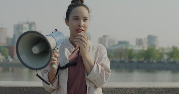 Portrait of young lady activist speaking in megaphone standing outdoors in river embankment in city. Social protest and human rights protection concept.
