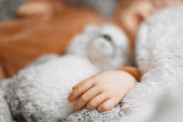 Close up hand of little baby lying in bed with fluffy stuffed toys of animals. Toddler sleeping with teddy bear in cozy room and enjoy healthy night sleep. Selective focus. 