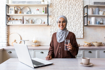 Young Muslim woman in hijab sitting at home in the kitchen with a laptop and holding a glass of pure mineral water. Leads a healthy lifestyle, smiles and looks into the camera, advertises, poses.