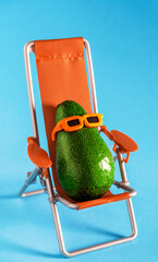 Cute avocado wearing sunglasses and sitting in beach lounger on a blue background. Summer minimal...
