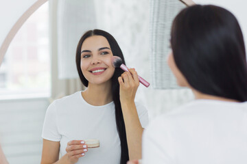 Smiling young beautiful woman stands in front of the mirror at home and applies make-up on her face with cosmetic bones.