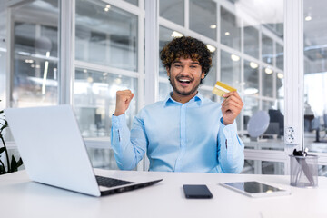 Young hispanic man sitting in the office at the desk, holding a credit card Looking at the camera and happy. Shows a victory gesture of success with his hands.