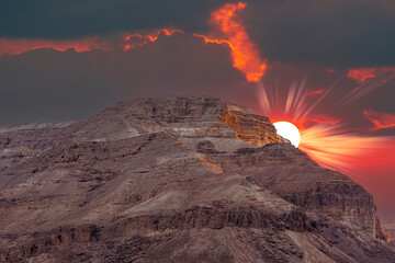 Sunset against backdrop of mountains and fiery red sun in the dark sky in Dead Sea, Israel. Beautiful desert landscape among the sands and stones. Large stone ridge with dramatic clouds and sun rays