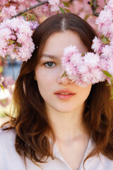 Naklejka premium Portrait of Young Caucasian Woman with Natural Beauty Posing Against Blooming Sakura Tree Outdoors. Dark Haired Lady Smiling and Looking at Camera