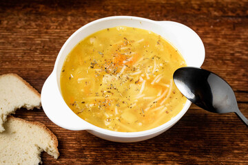 Chicken soup with noodles, potatoes, onions and carrots in a white deep plate with a black spoon and white bread on a wooden background.
