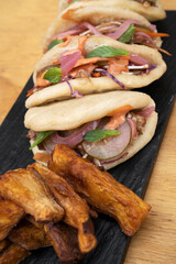 Asian cuisine. Closeup view of pulled pork baos buns with pickles, sauce and fried sweet potatoes, in a black dish on the wooden table.