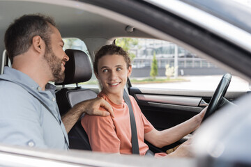 cheerful father putting hand on shoulder of teenage son while teaching him how to drive car.