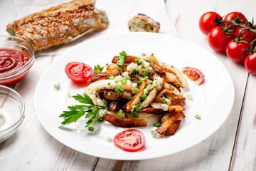 Fried potatoes in a white plate with green onions, parsley, tomatoes on a white wooden background.