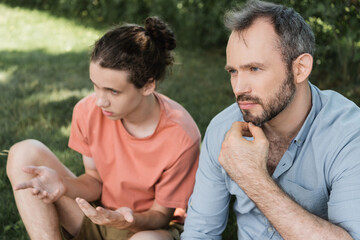 pensive and bearded father sitting next to teenage son while having conversation in green park.
