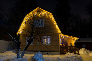 Yellow Christmas lights garlands hangs on wooden residential house at night. Soft focus. Holidays...