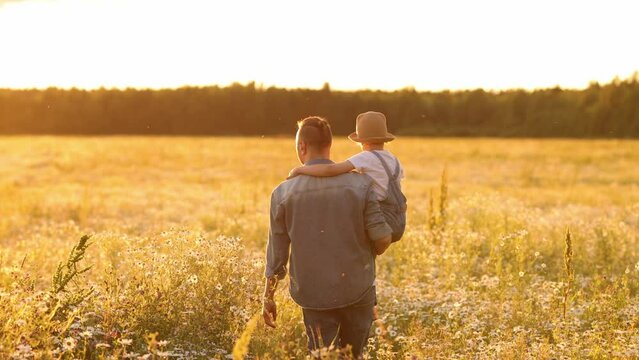 A young dad carries a little daughter in a straw hat in her arms. Walking through the chamomile field at sunset. The concept of life outside the city and walks outside the city on the weekend.