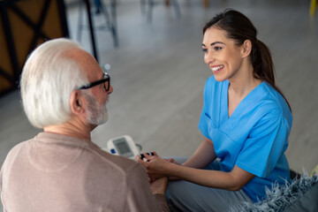 Lovely young nurse smiling and looking at older senior man during home visit check up. Home care,...
