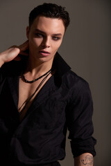 Obraz premium Studio portrait of a young teenager brunette boy in a black shirt with beads around his neck. Short trendy haircut. Elegant sexy style. Gray background.