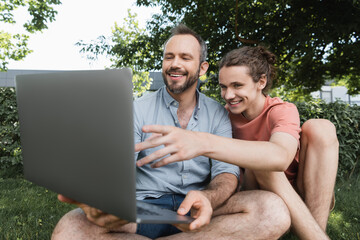 smiling father using laptop next to happy teenage boy while sitting together on green lawn.