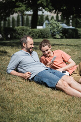 Fototapeta premium bearded man smiling while using smartphone and resting on lawn near teenage son after playing football.