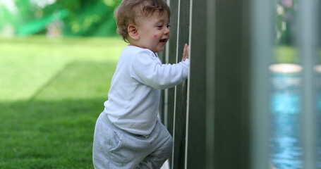 Infant baby holding into swimming pool fence. Toddler child standing by security protection outside in backyard