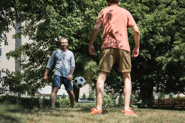 bearded man playing soccer with his teenage son in green summer park.
