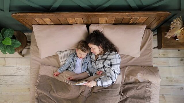 Top view of young woman reads a book with fairy tales to her toddler son before going to bed and kiss him. The child reads a book with his mother lying in bed. Happy childhood. Slow motion video