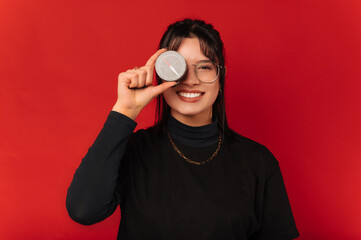 Cheerful young smiling woman is covering one eye with a black compass over red background.