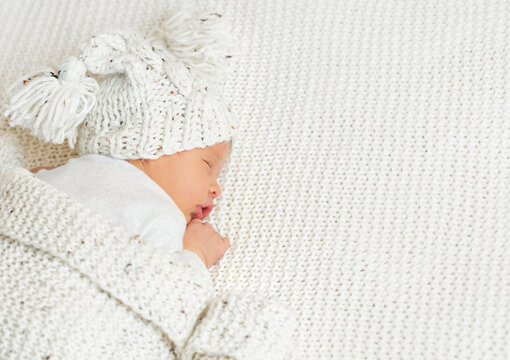 Baby In Woolen Hat With Pompon Sleeping On Knitted White Blanket. Newborn Lying Down On Stomach. Infant Child Health Care And Development