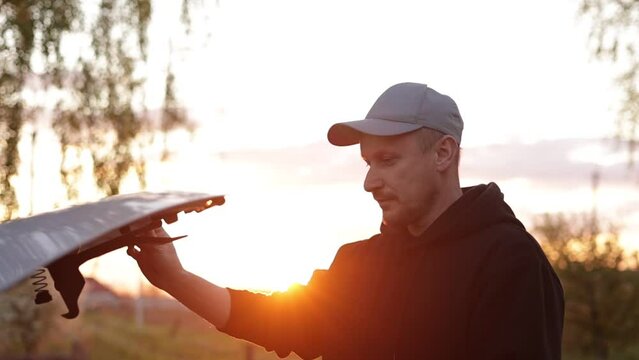 Young man is opening the hood of a car outdoors in the evening on sunset. Driver checking or fixing the automobile engine on the road after a car breakdown. Vehicle service concept. Slow motion.