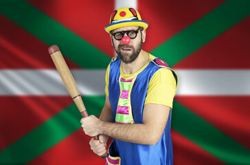 An insecure clown holds a bat in his hands against the background of the flag of Basque lands
