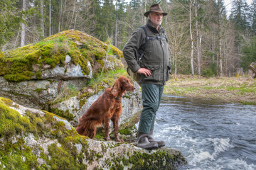 An elderly hiker and his trusty Irish Setter stand on a large rock jutting out of the water. Both...