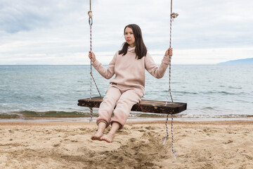 Happy young woman traveler on a wooden swing, a beautiful blue lake Baikal with waves, a sandy beach. Summer on Lake Baikal.A girl rides on a swing on the seashore. Travel