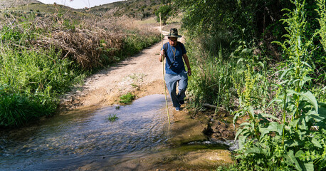 Obraz premium Rural dirt road with shallow creek crossing, man walking through water, lush green countryside travel landscape