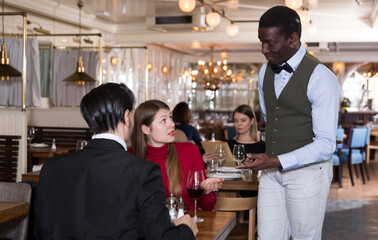 Hospitable African American waiter talking with young couple visiting restaurant