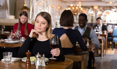 Pensive young woman sitting alone in restaurant table with glass of red wine