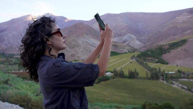 joven mujer latina de perfil grabando o tomando fotos con su celular al paisaje al atardecer en el Valle del Elqui Paihuano