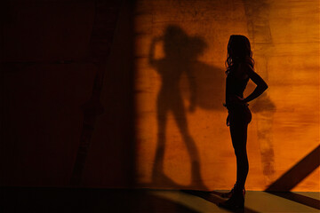 Woman from behind flexing muscles. To the right of the wall the shadow of the woman as a superhero with a cape