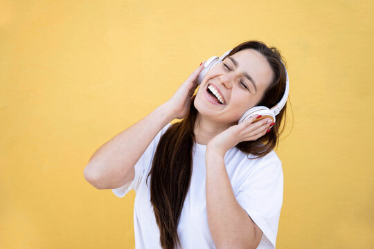 Happy young woman singing and having fun while listening music using wireless headphones - Cheerful girl posing isolated on pastel yellow wall background studio portait