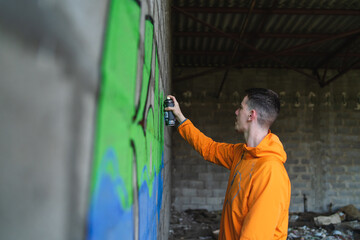 One young guy drawing graffiti on wall with spray can and mask on his face during the day