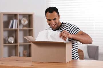 Happy young man opening parcel at table indoors. Internet shopping