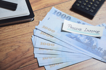 Passive income handwritten note on New Taiwan dollar banknotes with calculator on desk. Extra...