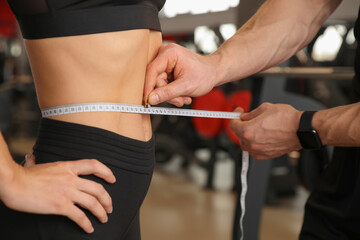 Fototapeta na wymiar Trainer measuring woman`s waist with tape in gym, closeup