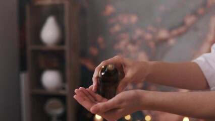 The cosmetologist or massage therapist in the beauty salon's room holds a dark jar of beauty treatment fluid and pours it onto their hands in the air.