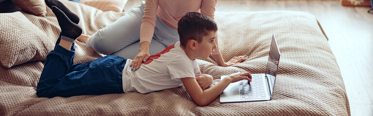 Beautiful young Caucasian mother sitting on bed and watching small son using laptop.